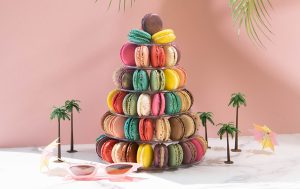 A French macaron pyramid with assorted flavors surrounded small palm trees, a pair of sunglasses, and two cocktail umbrellas.