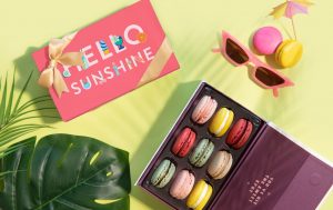 A box full of assorted macarons surrounded by a Woops! Macaron boxes, a pair of sunglasses, a cocktail umbrella, and palm leaves.