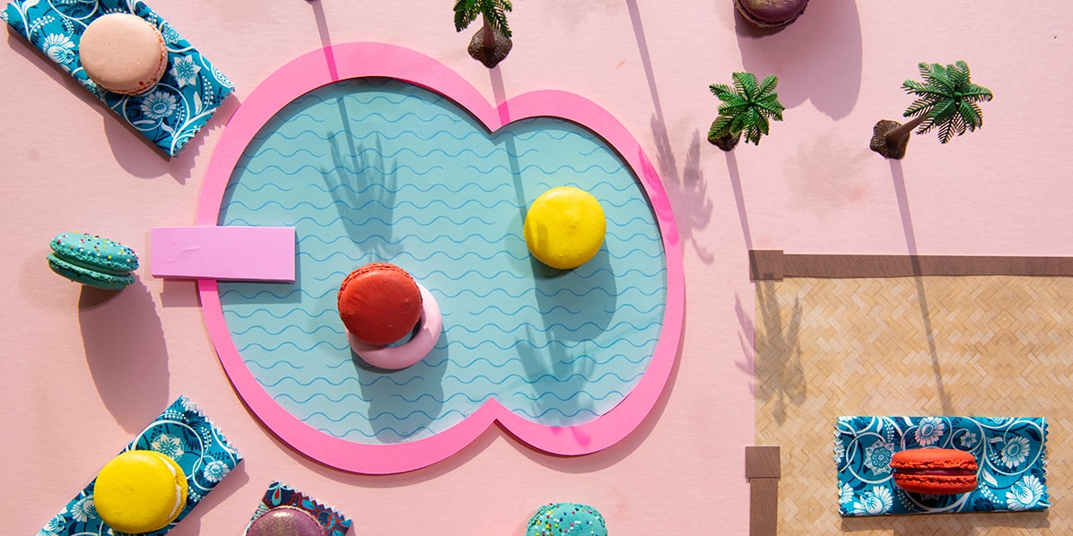 Assorted macarons and small palm trees are surrounding a cardboard pool.