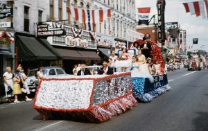 A blue, white, and red decorated card full of people parading with people watching. Above are some blue, white, and red flags.