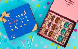 A box full of 18 assorted macarons has a macaron box with a dark blue Birthday sleeve to the left. Surrounding them is confetti and below tiny candles.