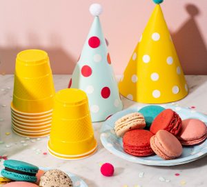 Two green Woops! Macaron boxes with candles on top are surrounded by assorted macarons, party hats, and yellow cups. 