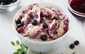 A bowl full of coconut and berry ice cream surrounded by blueberries and a tiny bowl with berry jam. 