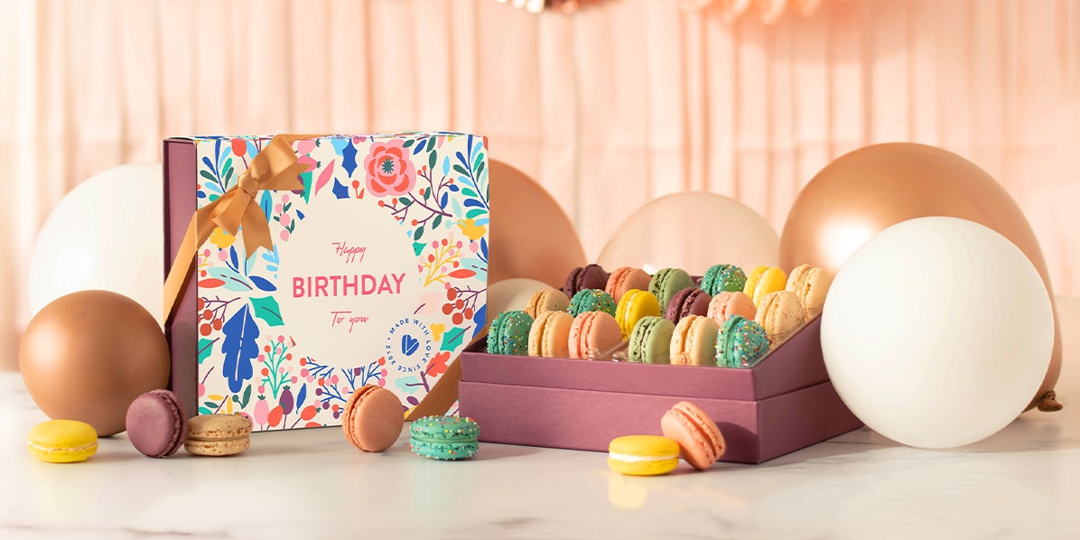 A box full of assorted macarons has a Woops! Macaron box with a Birthday sleeve to the left. Surrounding them are balloons, party decorations, and macarons.