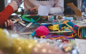 Numerous crafting items on a table and a woman with a sweater sitting on the background.