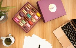 A box full of 18 assorted French macarons surrounded by a computer, paper, a cup of tea, and a tiny plant.