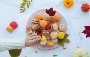 A female hand is grabbing a macaron from a plate full of assorted macarons and leaves. Surrounding the plate are some macarons,