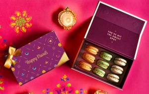 A box full of 9 assorted macarons is surrounded by a Diwali sleeve, confetti, and Diwali candles.