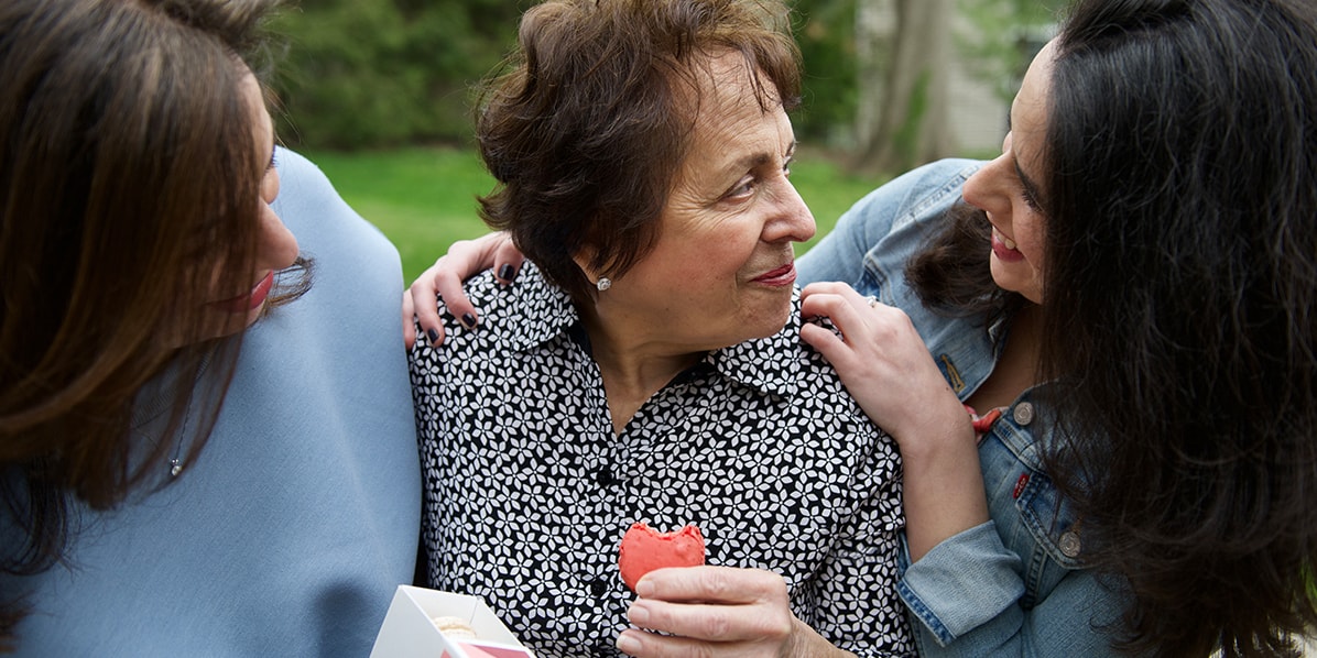 Three smiling women are looking at each. The older woman in the middle is holding a macaron in her hand and the one to the left is holding a Woops! Favor box of macarons in her hand.