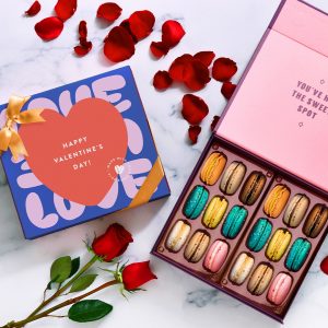 An opened box of 18 French macarons with assorted flavors has a box of 18 French macarons with a Valentine's Day sleeve to its left. At the bottom are 2 red roses and above some red rose petals.