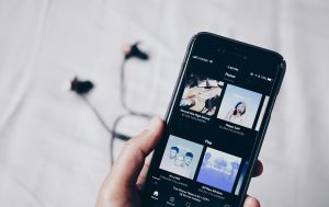 Hand holding a smartphone displaying Spotify playlists, with earphones resting on a white surface.