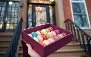Hand holding a box of colorful Woops! macarons in front of a brownstone building entrance.