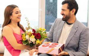 Smiling couple exchanging flowers and a Woops! macaron box labeled 'LOVE' while sitting on a couch.