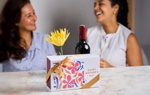 Two women, with a box of macarons, wine and a flower on the table in front of them.