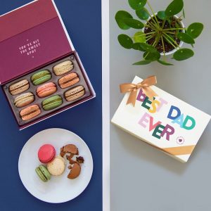 A box full of nine assorted macarons has a plate full of macarons below and a macaron box with a Father's Day sleeve plus a plant to the right.