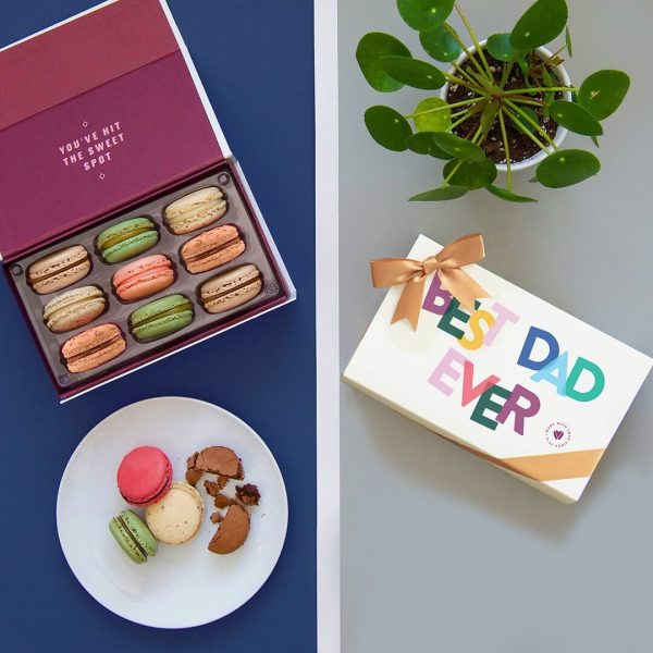 A box full of nine assorted macarons has a plate full of macarons below and a macaron box with a Father's Day sleeve plus a plant to the right.