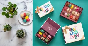 Flat lay of colorful macaron gift boxes for Father's Day, including boxes labeled 'Best Dad Ever' tied with gold ribbons. Some macarons are arranged on a white plate surrounded by green houseplants on a marble and teal background.