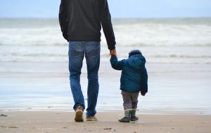 Man and boy walking on seashore under blue sky.
