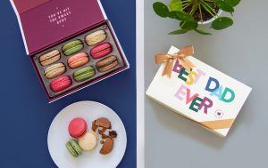 An opened box of macarons in the left with 4 macarons in a plate and an box of “Best Dad ever” in the right. 