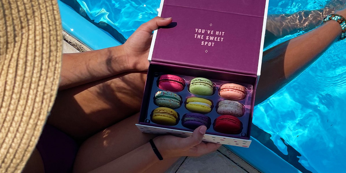 Woman with a hat has her legs inside the pool while holding an open box of Woops! macarons.