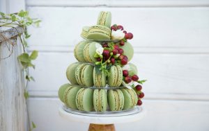 A Woops! macaron pyramid decorated with red grapes and white flowers, displayed on a marble cake stand.