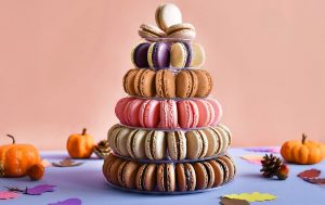Colorful macaron tower arranged in a pyramid shape with fall decorations, mini pumpkins, and paper leaves on a pastel background.