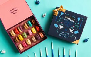 Open Woops! macaron box with colorful macarons beside a closed Hanukkah-themed gift box decorated with a menorah and gold bow, surrounded by blue candles and dreidels on a turquoise background.