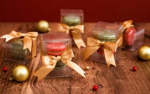 Small clear macaron boxes tied with gold ribbons arranged on a wood surface, surrounded by holiday ornaments, greenery, and red berries.