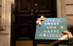 Hand holding a Woops! holiday gift box with a gold ribbon that reads “Have a Sweet and Happy Holiday!” in front of a dark wooden door.