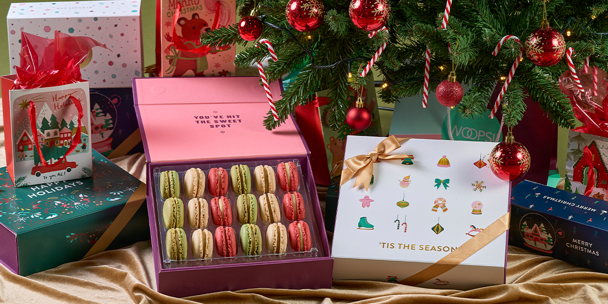 Colorful Woops! macaron gift boxes and holiday bags arranged under a decorated Christmas tree with red ornaments, candy canes, and lights on a gold fabric backdrop.