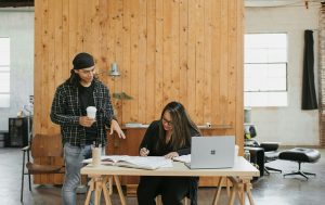 A man standing next to a woman working on a laptop.