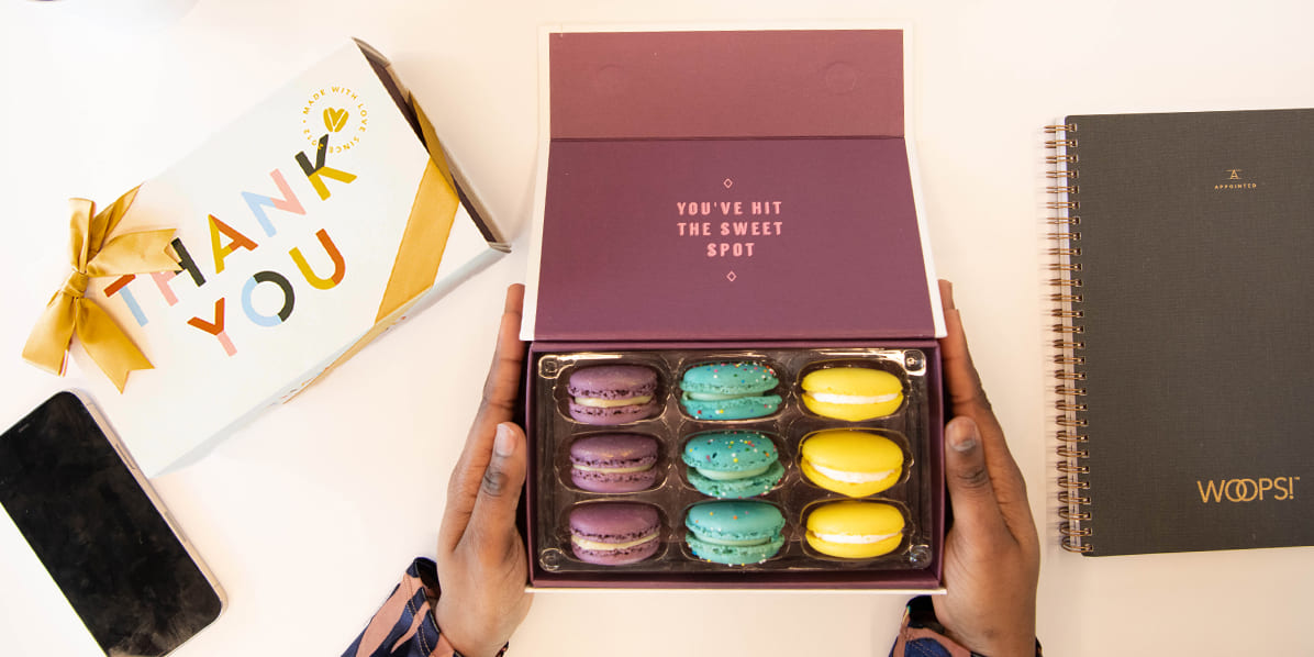 Hands holding a Woops! macaron gift box with colorful macarons on a desk, alongside a “Thank You” box, notebook, phone, and office supplies.