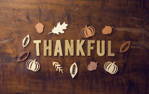 The word “THANKFUL” in glittery gold letters surrounded by wooden cutouts of leaves, acorns, and pumpkins on a wooden table.