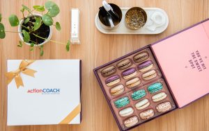 Open Woops! macaron box with assorted macarons beside a branded ActionCOACH gift box on a wooden desk with office supplies and a small potted plant.