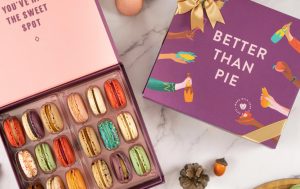 Open Woops! macaron box with colorful macarons beside a closed purple “Better Than Pie” gift box tied with a gold ribbon, surrounded by fall accents and extra macarons on a marble surface.
