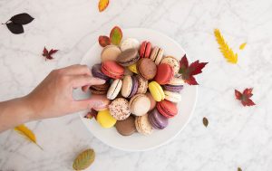 A female hand is grabbing a macaron from a plate full of assorted macarons and leaves. Surrounding the plate are some fall leaves