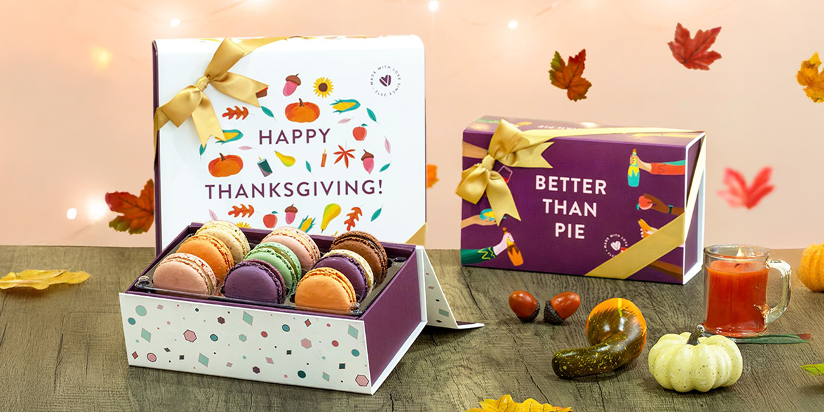 Woops! Thanksgiving macaron gift boxes with gold ribbons displayed on a wooden table surrounded by fall leaves, pumpkins, and acorns, with glowing lights in the background.