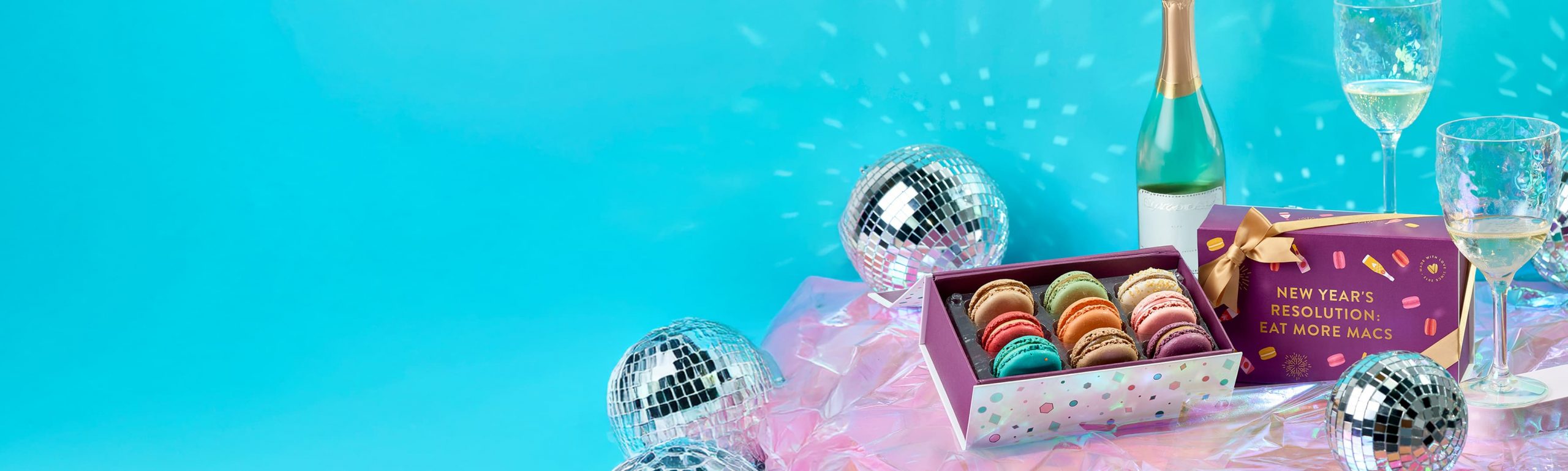 Woops! New Year’s macaron gift boxes with gold ribbons displayed on a shiny surface surrounded by disco balls, champagne glasses, and festive decorations.