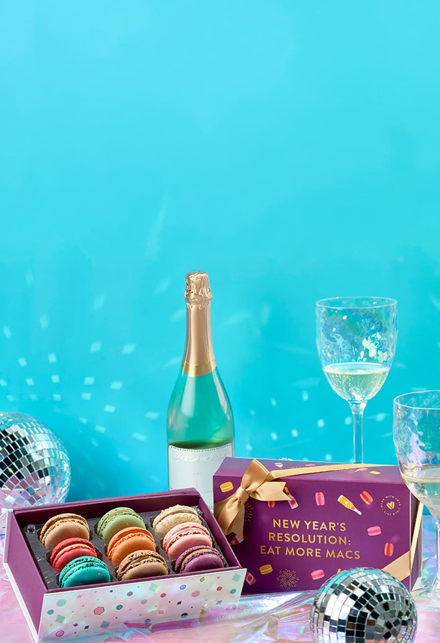 Woops! New Year’s macaron gift boxes with gold ribbons displayed on a shiny surface surrounded by disco balls, champagne glasses, and festive decorations.