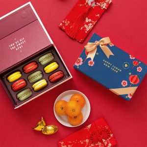 Lunar New Year macaron gift set with an open box of colorful macarons, mandarins, gold ingots, and festive red envelopes on a red background.