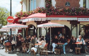 Busy Parisian café with red awnings and flower-covered façade in Montmartre, as people sit at small outdoor tables under white umbrellas on a sunny day.