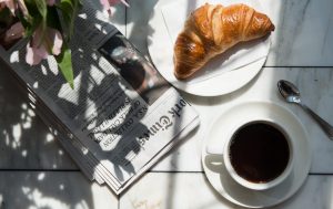 Fresh croissant on a white plate beside a cup of black coffee and a folded newspaper on a marble table, with soft sunlight and floral shadows creating a cozy café atmosphere.