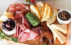 Charcuterie board with sliced salami and prosciutto, assorted cheeses, olives, crusty baguette slices, herb-coated cheese, fig spread, and dried sausage on a wooden serving board.