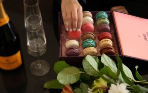 Hand reaching into an open box of assorted colorful macarons beside two champagne flutes and a bottle of Veuve Clicquot, with fresh flowers on a dark table.
