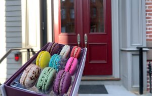 Hand holding an open box of colorful assorted macarons in front of a red double-door entrance to a residential building.