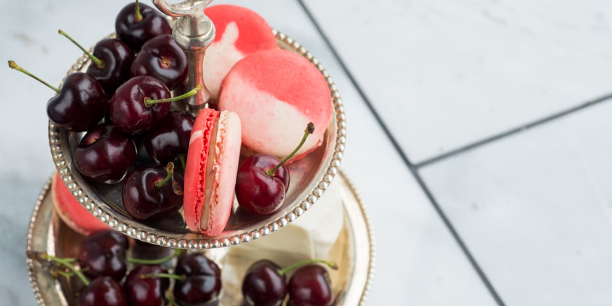 Two-tier silver dessert stand filled with fresh cherries and pink macarons, set on a light tiled surface.