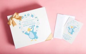 White gift box with gold ribbon featuring a blue parrot illustration and invitation text, displayed on a pink background alongside a matching printed event card and envelope.