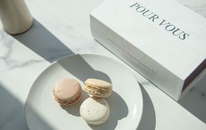 Three neutral-toned macarons on a white plate beside a minimalist white “POUR VOUS” gift box on a marble surface with soft natural light and shadows.
