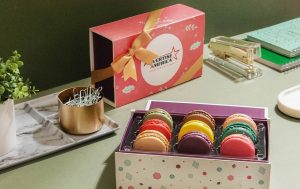 Open box of assorted colorful macarons on a green desk beside office supplies and a branded gift box tied with a gold ribbon against a dark green wall.