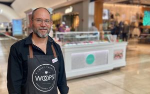 Smiling man wearing a black Woops! apron standing in a shopping mall in front of a macaron kiosk display. 